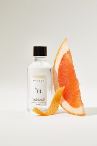 sunday studio grapefruit peel oil polish remover with a slice of grapefruit leaned on the bottle and a grapefruit peel in front of the bottle