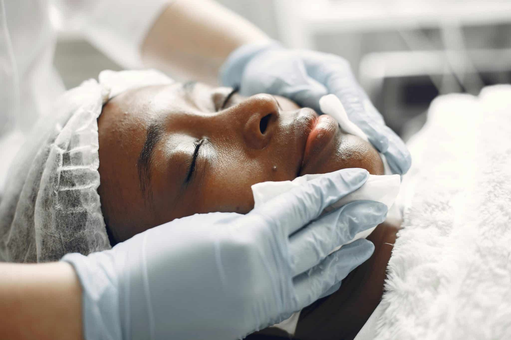Close-up of a woman’s face during a facial treatment, wearing a white hair cap while an esthetician in blue gloves performs the procedure.