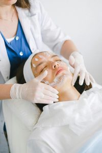 Esthetician in a white jacket and blue blouse gently massages a woman's face with facial cream. The client, wearing a white headband and towel, lies on a white massage table with eyes closed in a bright, white-walled spa room.