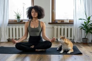 Woman meditating on yoga mat in an apartment next to her dog.