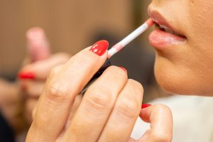 close-up of a white woman, with red painted nails, applying lip gloss
