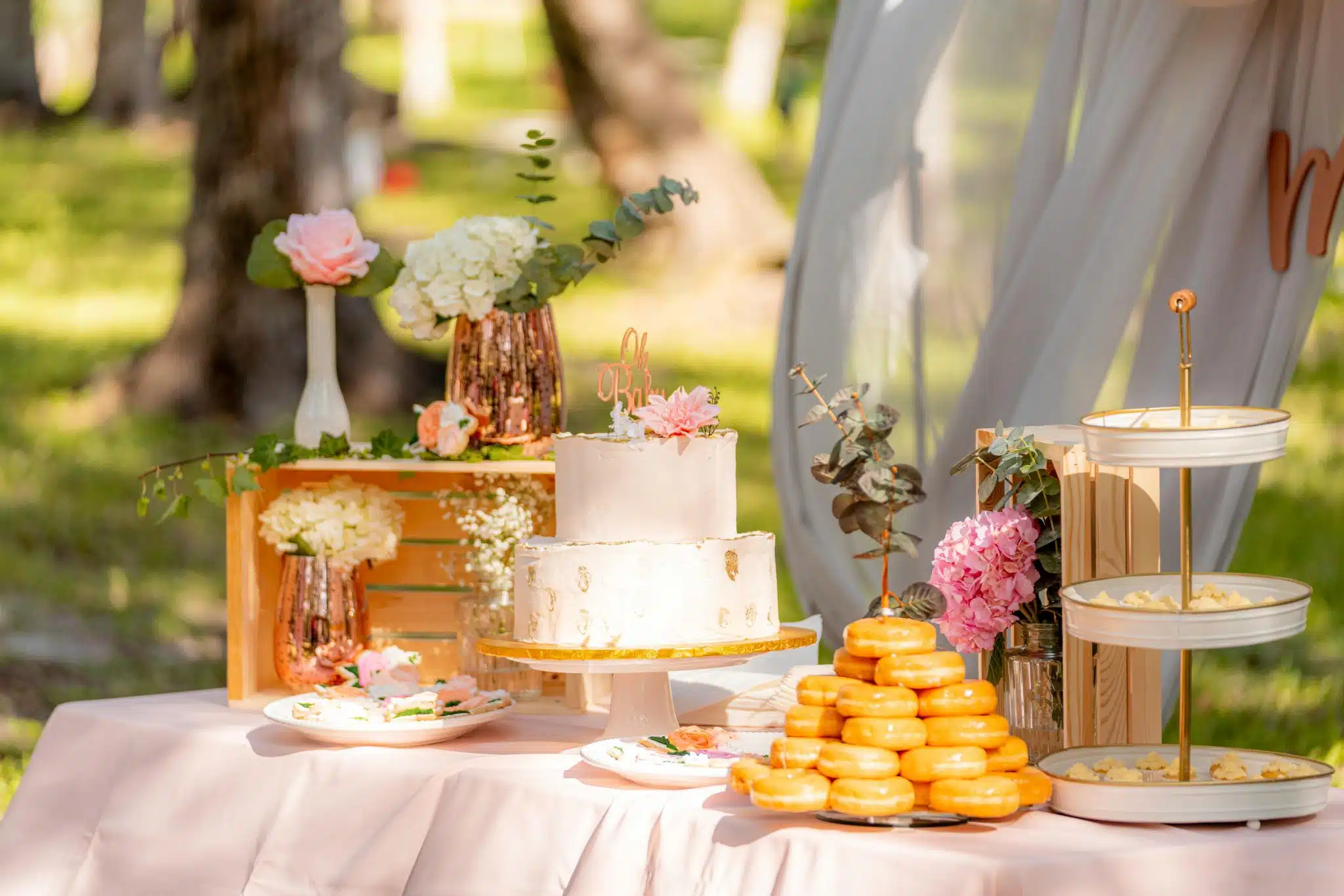 Outdoor table with a pink tablecloth, topped with a cake, a tower of donuts, and flowers; sheer drapery billows in the wind in the background