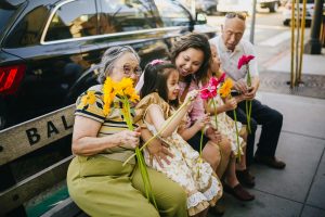 asian family sitting on a street bench in front of a black car, in order from left to right starting with grandmother, grandchild, mother, grandfather