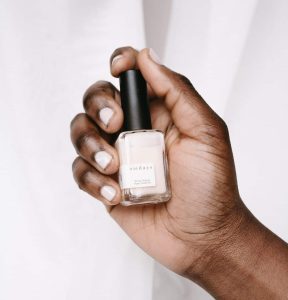 close-up of a black woman's hand, with neutral polished nails, holding a sundays studio nail polish bottle