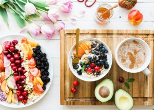 countertop with a bowl of cut fruit next to a wood cutting board with an avocado that has been cut open, hot beverage in a mug, and bowl of yogurt with fruits and nuts. there are alos flowers, honey and other produce on the countertop