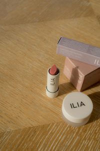 ILIA uncapped lipstick and a product in a circular jar on a wooden table.