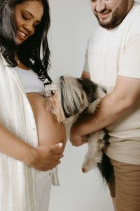 Expecting parents dressed in white smiling in front of a white wall, as their dog playfully licks the woman’s baby bump.