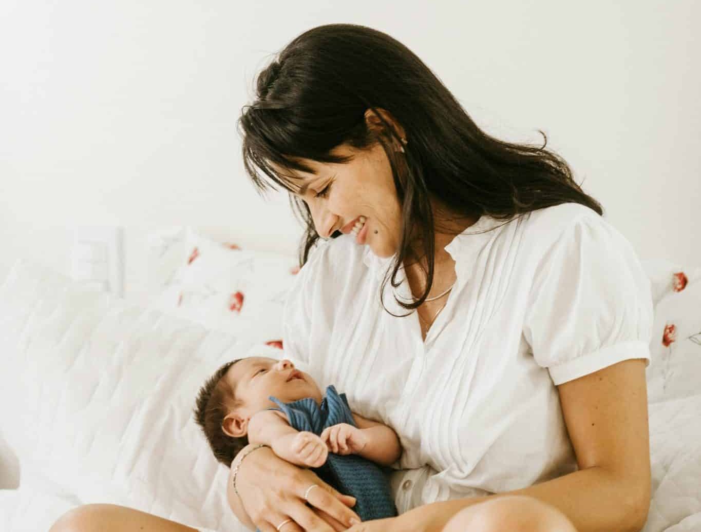A mother in a white linen set sitting on white bedding, holding her newborn baby