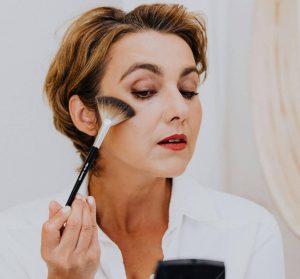 Woman in a white button down against a white background applying makeup with a fan brush while looking in a compact mirror.