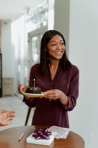Smiling woman in a dark burgundy dress holding a chocolate birthday cake with a lit candle, surrounded by friends in a sunlit room with white walls. A wooden table with cards and a gift is in the background.