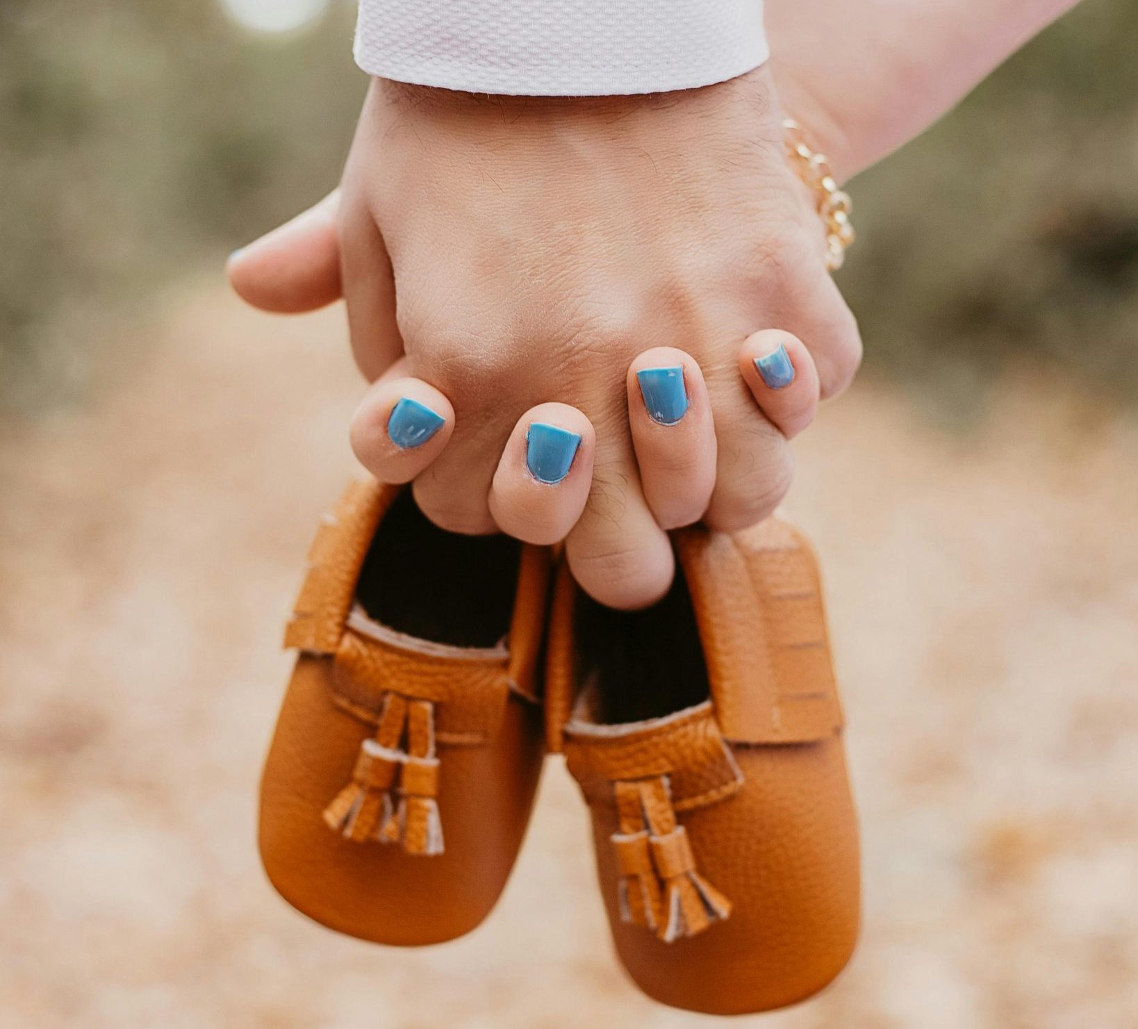 Close-up of a man and woman holding hands while carrying baby shoes; the woman wears soft blue nail polish, symbolizing they are expecting a baby boy.