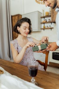 Woman in a purple sundress sitting at a wooden kitchen table, unwrapping a green birthday gift with a pink ribbon. She has black nail polish and a joyful expression. There is a glass of wine on the table.