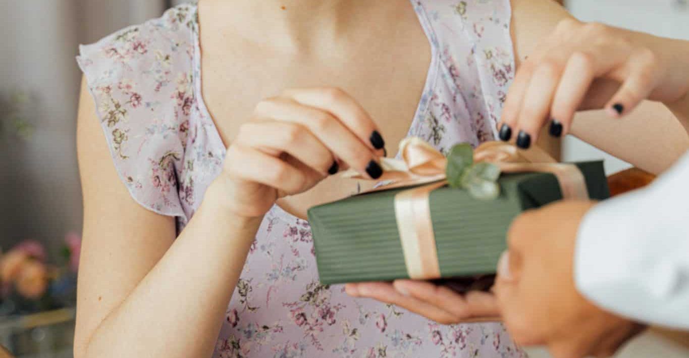 Woman in a purple sundress sitting at a kitchen table, unwrapping a green birthday gift with a pink ribbon. She has black nail polish and a joyful expression.