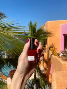 Women holding a red nail polish bottle that matches her finger nails in front of an hacienda with a tropical view.