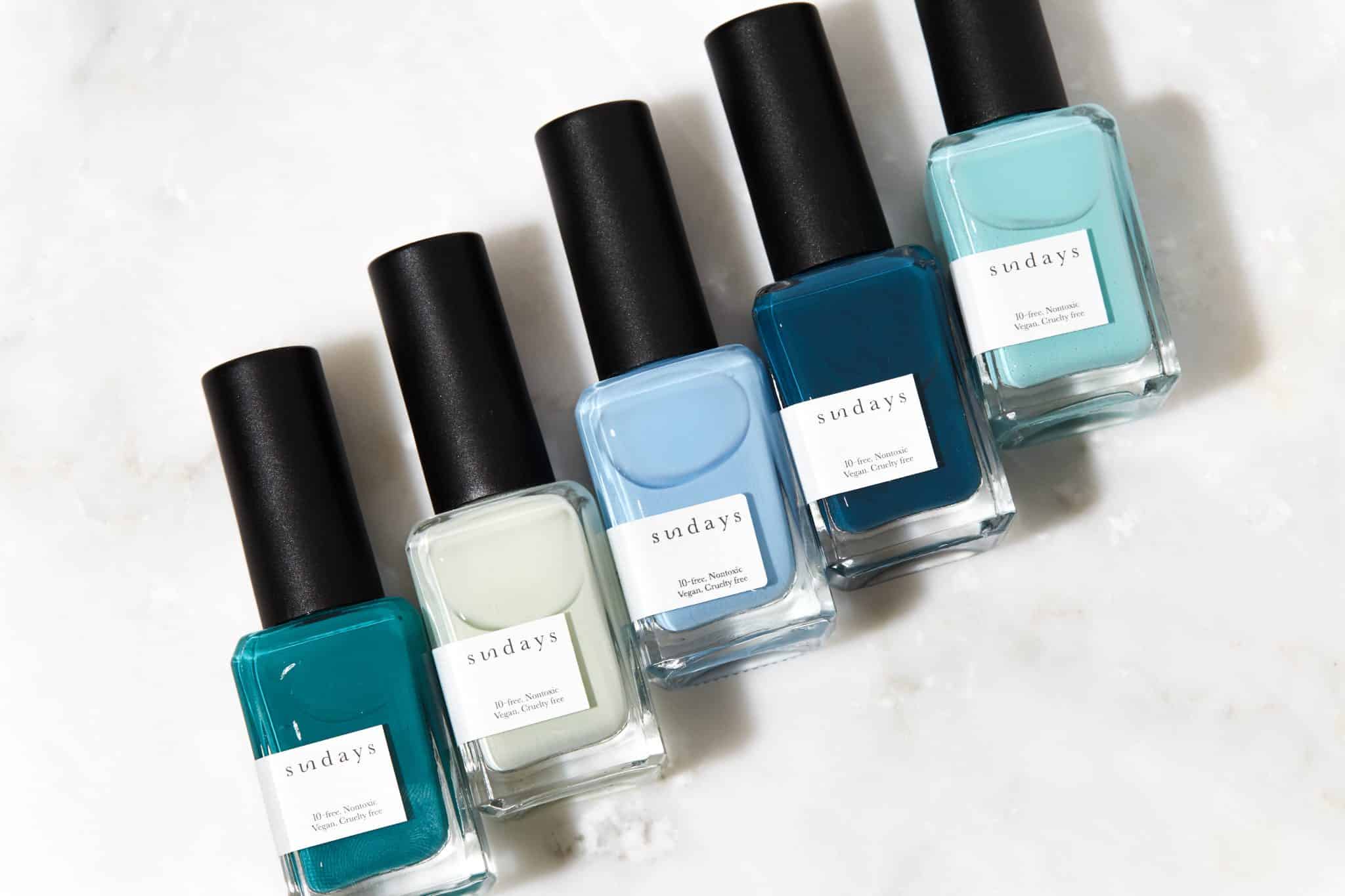 5 cool-toned blue and green nail polish bottles lying on a marble countertop.