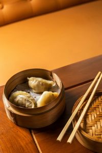 3 dumplings in a bamboo steamer with chopsticks resting on the steamer lid on top of a wooden table
