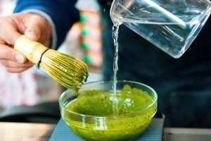 Close up of a man holding a matcha whisk in one hand and pouring hot water into a clear glass bowl with matcha powder with the other hand.