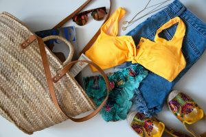 A woven tote bag, yellow bikini top, denim shorts, sunscreen, tennis shoes, sunglasses, a patterned bikini bottom, and other beach contents splayed on a white table.
