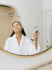 Woman in a white robe spraying her face with facial mist in the bathroom mirror.