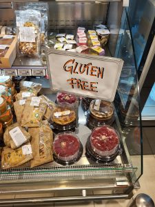 Refrigerated bakery case filled with gluten-free cookies, cakes, and assorted baked goods, with a handwritten 'Gluten Free' sign displayed prominently in bold marker.