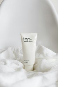 A white bottle of Necessaire body lotion on a white blanket with a white wall in the background.