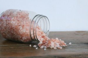 A glass jar of pink himalayan salt on its side, spilling onto a wood table.