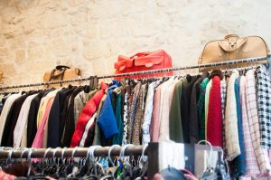 Clothing rack in a thrift store with long sleeve tops and large leather bags against a tan stone wall.