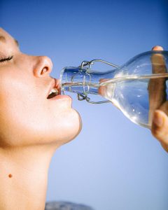 Woman drinking water from a glass bottle in front of a luminescent sky blue wall.