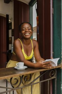 Woman wearing a yellow top and skirt smiling and reading in a wooden table sill at a coffee shop.
