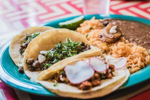 Three tacos garnished with fresh cilantro and radishes, served with Spanish rice and refried beans on a blue oval plate, placed beside a glass of water on a red-and-white chevron tablecloth.