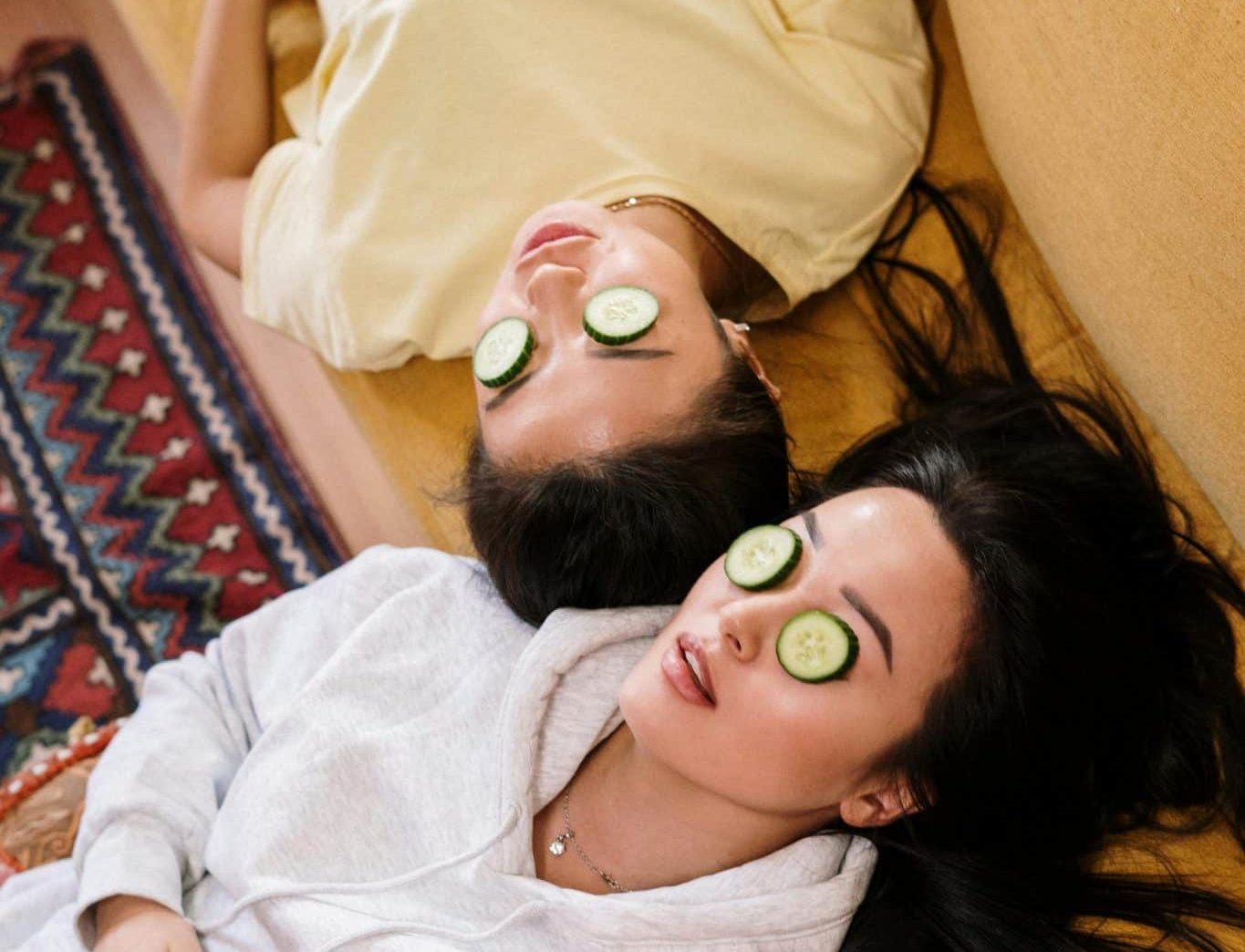 Two women laying on a couch in lounge clothes with cucumbers over their eyelids.