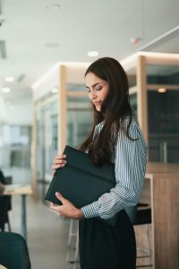 Woman standing in an office wearing a blue striped button up and a black pencil skirt, holding a black leather portfolio in her arm.