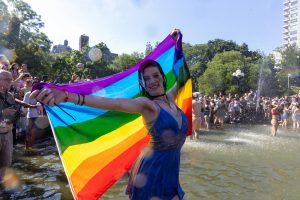 Woman holding pride flag behind her in Washington Square Park fountain.