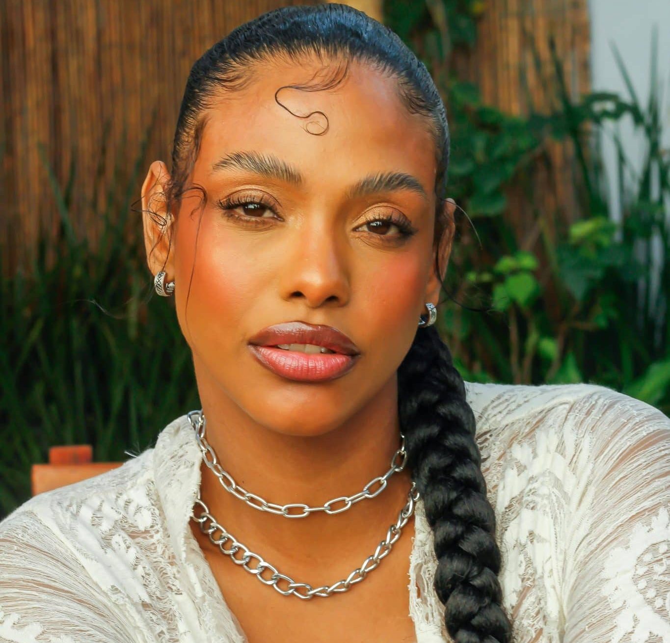 Woman with glowy, summermakeup wearing a silver chain necklace and white patterned blouse with a long slicked back hair braid.