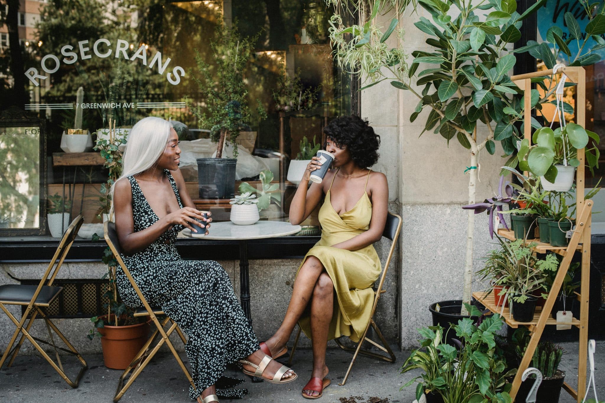 Two women wearing sun dresses and sandals sitting outside a coffee shop drinking beverages and chatting.