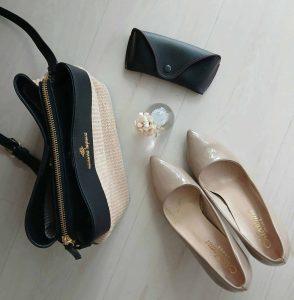 A women's purse, tan pumps, and sunglasses case arranged on a grey floor.