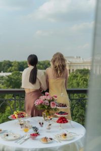 Rear view of two women, one in a yellow dress and one in a skirt and sweater, standing out on a balcony in front of a white table with various baked goods and flowers.